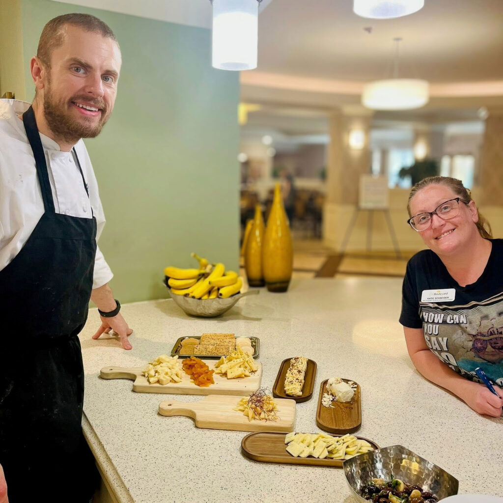 A chef and staff member stand by a counter with a charcuterie spread of cheeses, crackers, and dried fruit, showcasing creative dining offerings for residents.