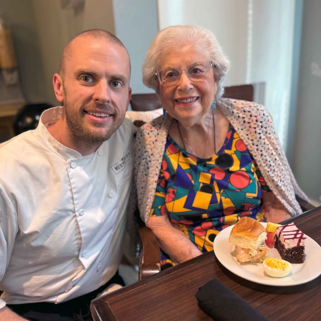 A chef smiles beside a senior resident enjoying lunch with a plate of sandwich, deviled egg, and dessert, highlighting dining experiences at the community.