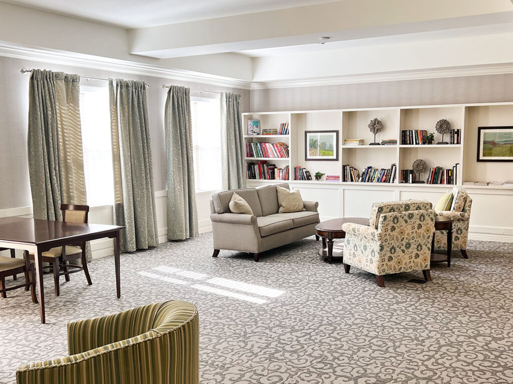 Library at Boulevard of Saint Charles Senior Living with cozy seating, bookshelves, large windows with curtains, and a table.