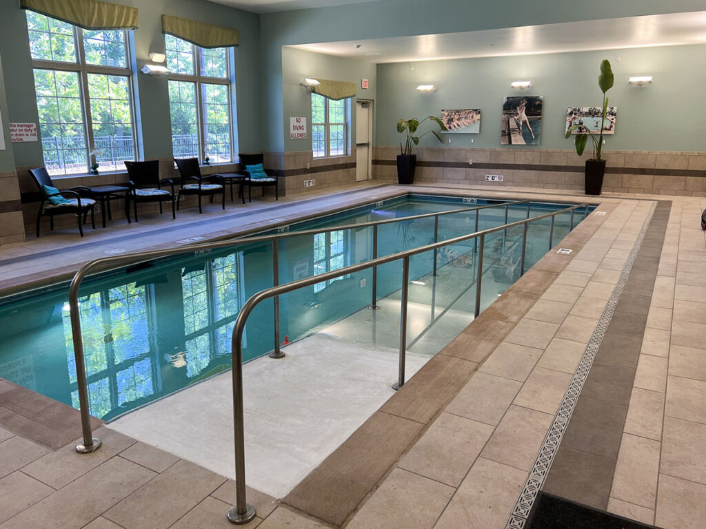 Indoor pool at the Boulevard of Saint Charles Senior Living, with handrails, seating area, large windows, and tiled flooring.