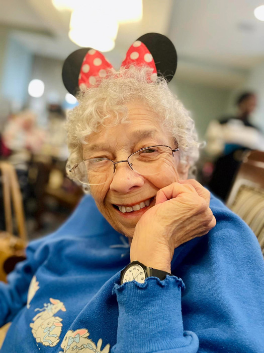 An elderly resident enjoys a Disney-themed happy hour, wearing red and black Minnie Mouse ears and a blue sweatshirt decorated with playful designs. She smiles warmly, resting her chin on her hand, adding to the cheerful ambiance of the event.