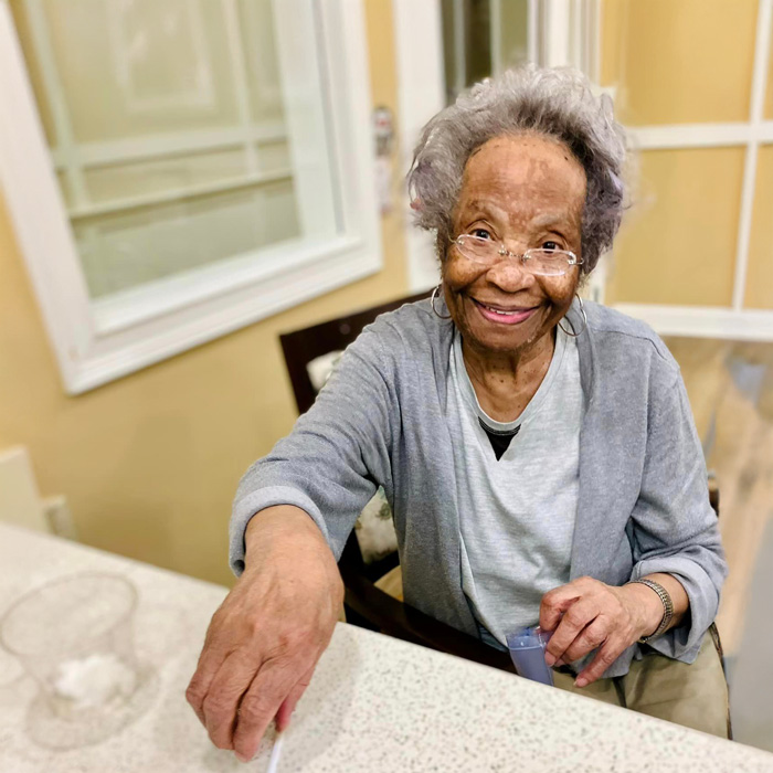 A smiling elderly resident enjoys a pop-up happy hour event, reaching out with a glass in hand. She is dressed in a light gray cardigan and a white top, sitting at a counter in a warm and inviting room