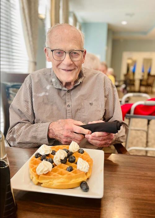 An elderly gentleman enjoys a delightful breakfast at a waffle bar, featuring a golden waffle topped with blueberries and whipped cream. He smiles warmly at the camera.