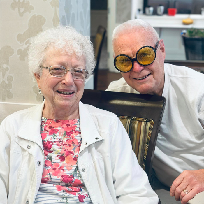 A couple enjoys a fun moment at the Don't Step on a Bee Day event. The woman wears a white jacket with a floral top, and the man sports humorous bee-themed glasses, creating a joyful and lighthearted atmosphere.