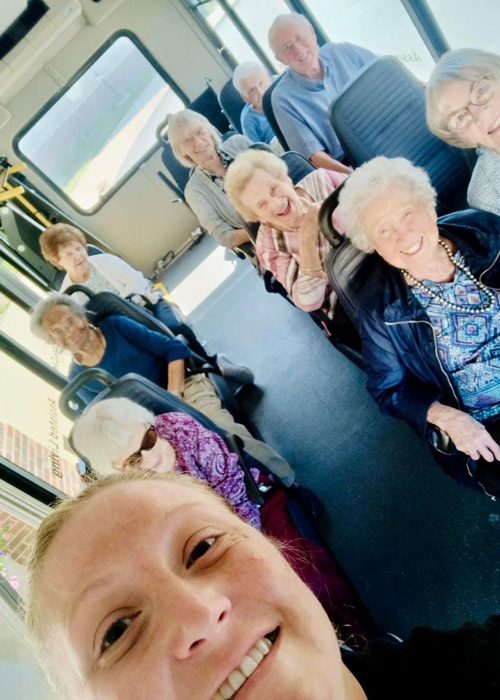 Group of smiling senior residents on a bus trip with a young woman taking a selfie in the foreground at a senior community.