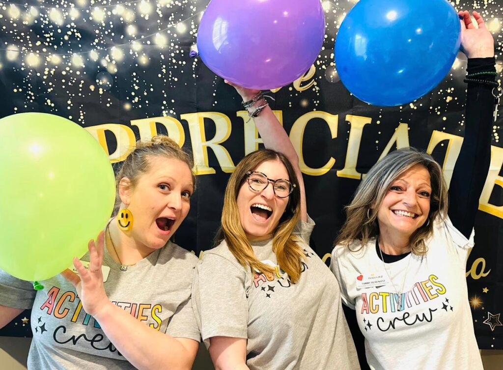 Three excited staff members from the "Activities Crew" pose with colorful balloons in front of a festive "Appreciate" banner decorated with twinkling lights during an employee appreciation event.