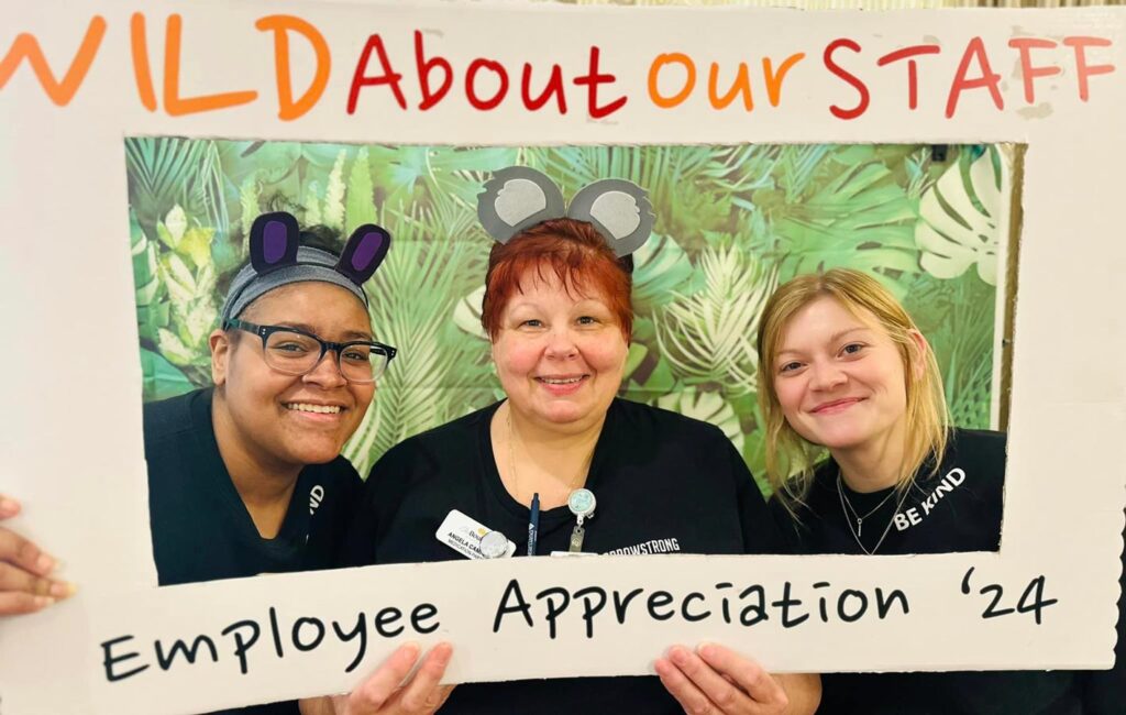 Three smiling staff members wearing animal ear headbands pose together behind a sign that reads "Wild About Our Staff, Employee Appreciation '24," against a tropical-themed background.