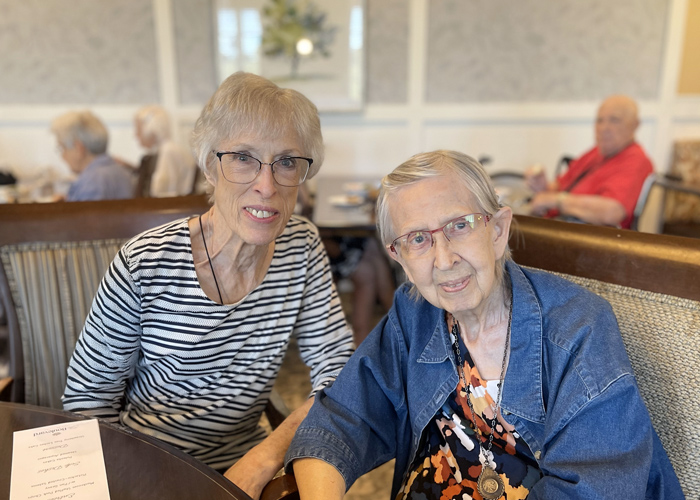 Two senior residents share a warm moment at a senior living community dining area, enjoying conversation and companionship over a meal.