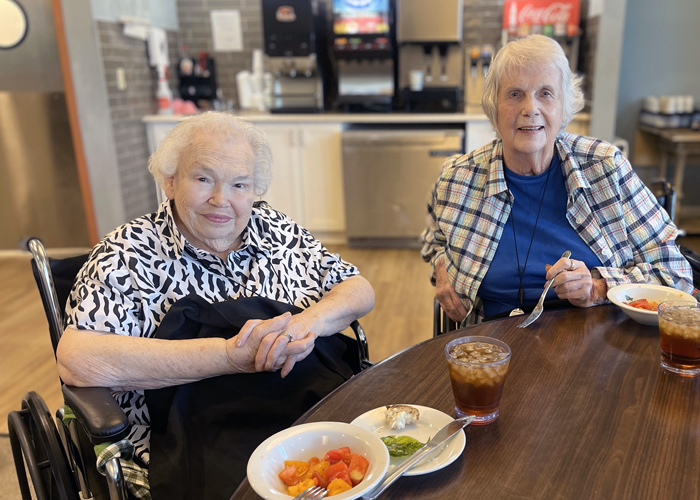 Two senior residents smile while dining together in a welcoming senior living community, enjoying fresh meals and meaningful connections.