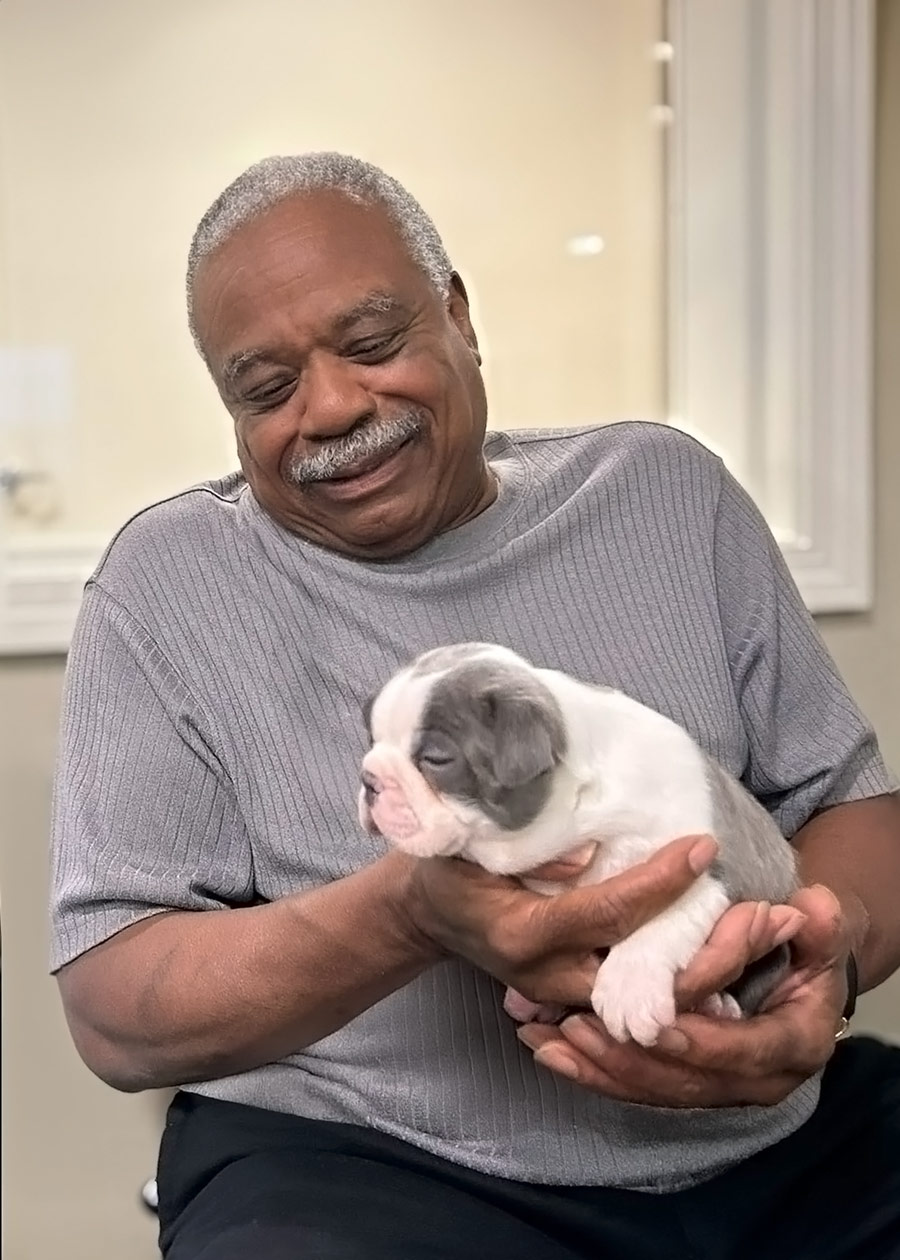 A senior resident with a warm smile, wearing a gray shirt, gently holds an adorable sleeping puppy in his hands, showcasing a moment of joy and affection.