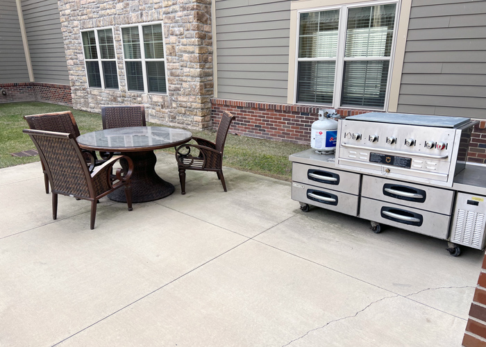 Outdoor patio with round table and four wicker chairs next to a large stainless steel grill station and propane tank outside a senior living community.