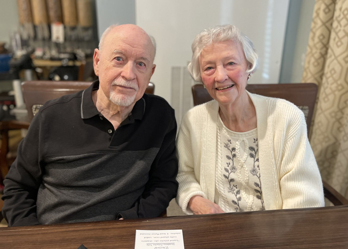 Senior couple sits together at a dining table, smiling warmly at the camera, enjoying quality time and connection during a community event or meal.