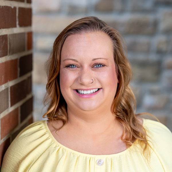 Natalie Miller, Senior Living Coordinator at The Boulevard Senior Living of Saint Charles, smiles brightly in a yellow blouse against a warm brick and stone backdrop.