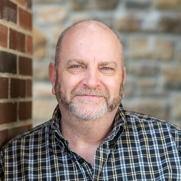 Paul Smith, Plant Operations Director at The Boulevard Senior Living of Saint Charles, stands against a brick and stone wall wearing a plaid shirt and a light smile.