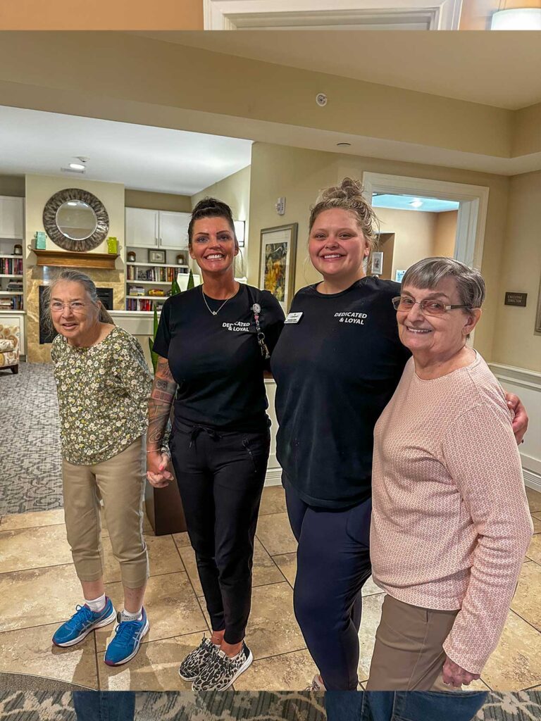 Two residents stand smiling with two team members in a bright community space, holding hands and showing connection, support, and joy in senior living.