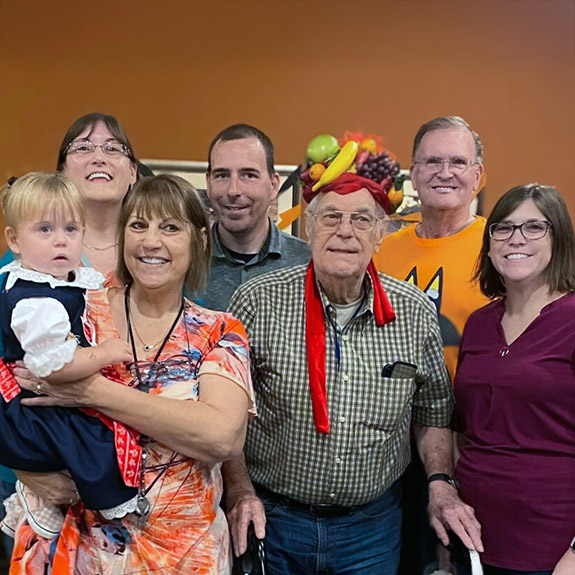 A senior resident wearing a novelty fruit basket hat smiles, surrounded by loved ones.