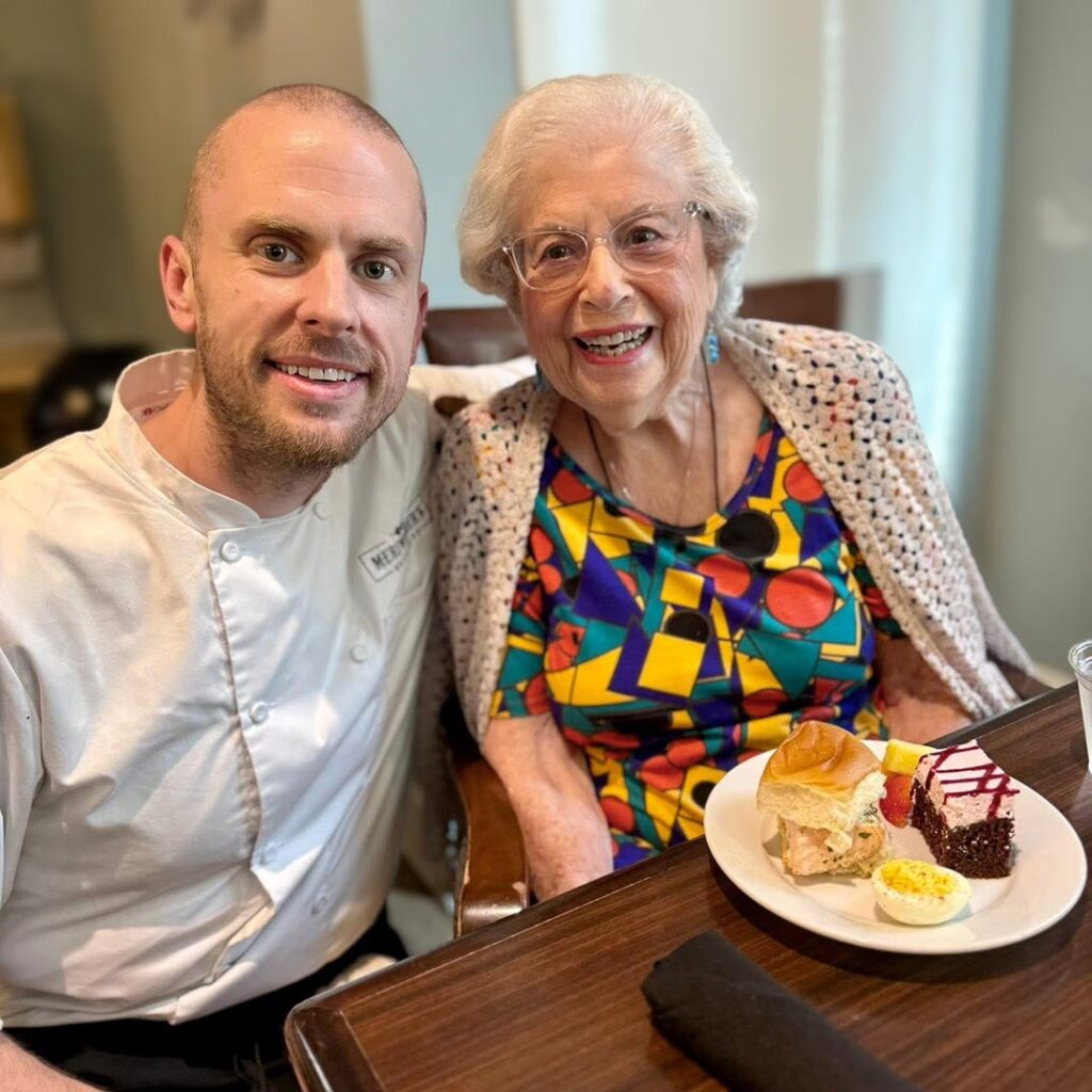 Culinary Director smiles alongside a senior woman with a plate full of food before her.