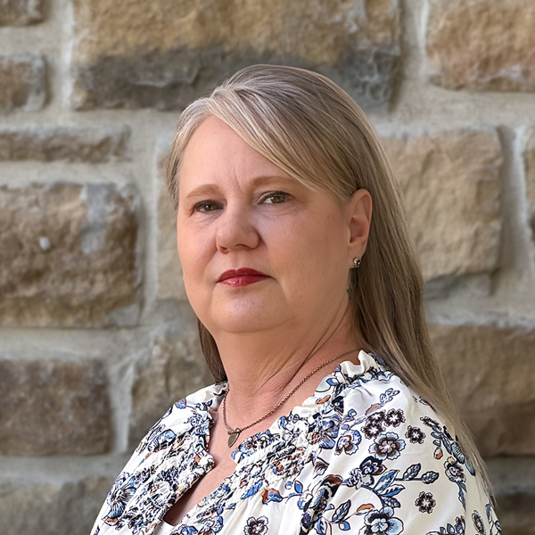Jennifer “Jen” Page, Wellness Director at The Boulevard Senior Living of St. Charles, looking at the camera in a professional headshot with straight blonde hair, a floral blouse, and a light stone wall background.