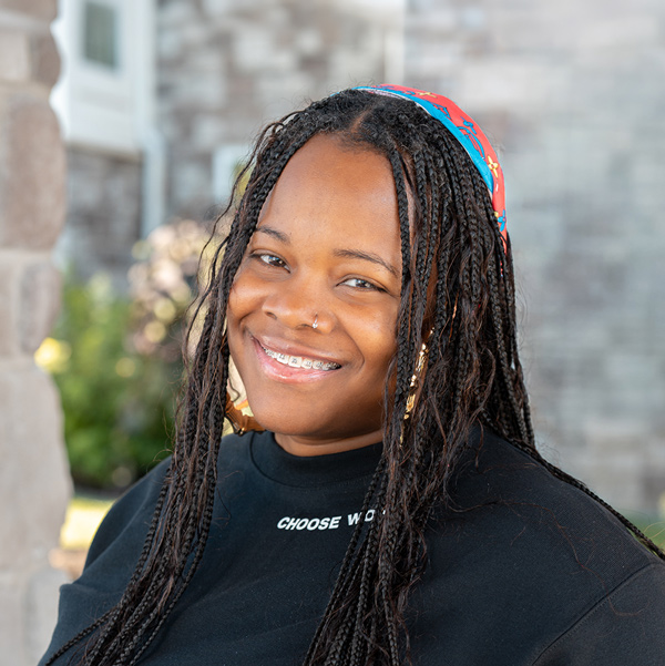 Brittany Crawford, Memory Care Director at The Boulevard Senior Living of St. Charles, smiling in a professional outdoor headshot with long braids, a colorful headband, a black top, and a softly blurred stone background.