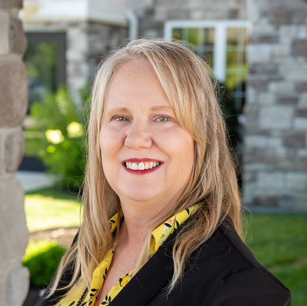 Jennifer Page, Wellness Director at The Boulevard Senior Living of St. Charles, smiling in a professional outdoor headshot with blonde hair, a black blazer, a yellow patterned blouse, and a softly blurred stone building background.