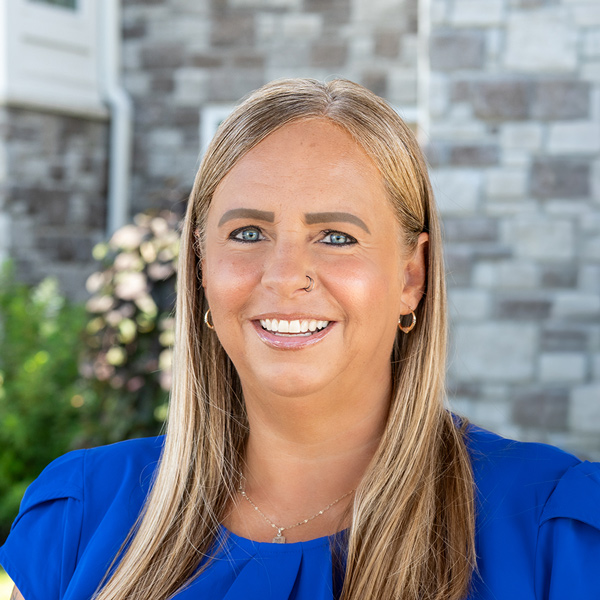 Melissa Schuff, Senior Living Coordinator at The Boulevard Senior Living of St. Charles, smiling in a professional outdoor headshot with long blonde hair, gold hoop earrings, a royal blue blouse, and a softly blurred stone building background.
