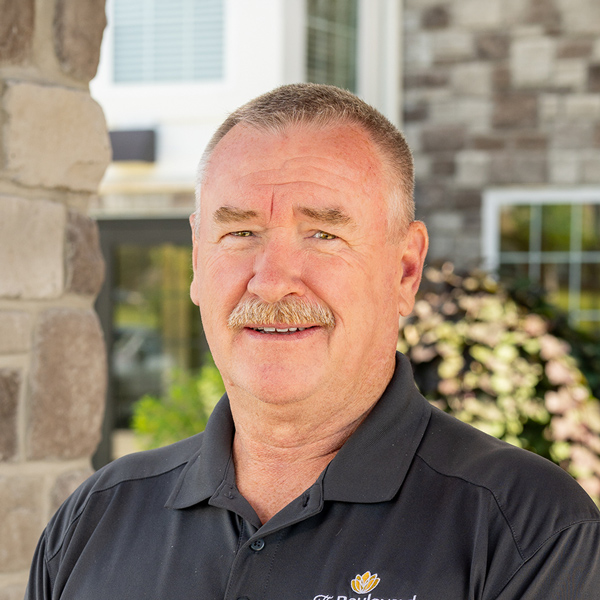 Robert “Bob” McKeon, Plant Operations Director at The Boulevard Senior Living of St. Charles, smiling in a professional outdoor headshot with short gray hair, a mustache, a black polo shirt, and a softly blurred stone building background.